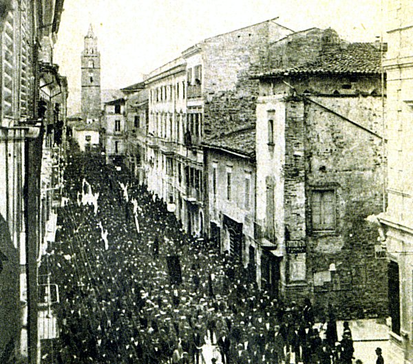 Immagine del palazzo nel 1920 (corteo del Primo Convegno provinciale dei lavoratori della terra). Visibile il palazzo Delfico di scorcio e lo stato della torre che, a quella data, appare in pessime condizioni di manutenzione